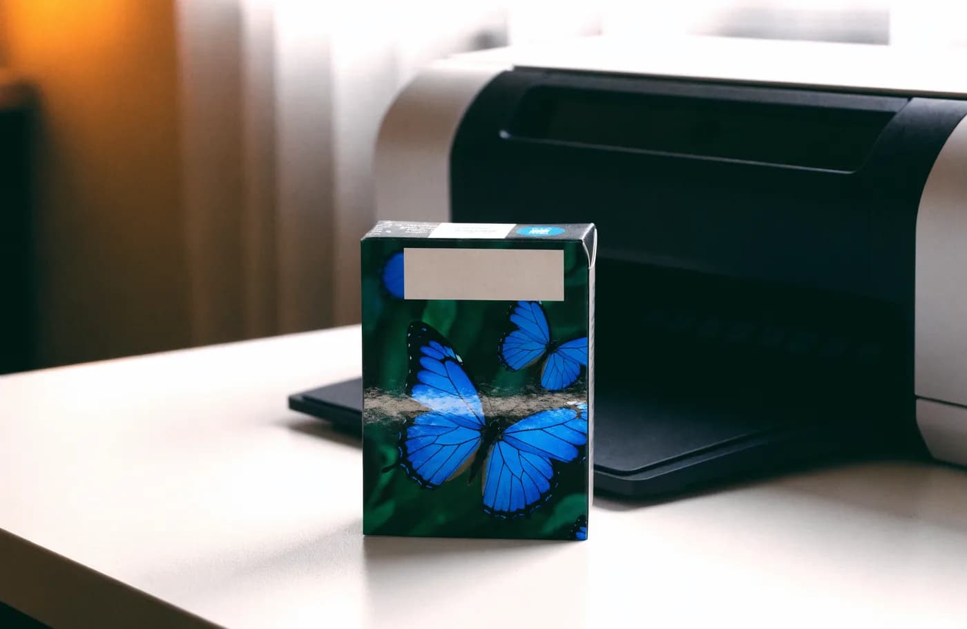 Boîte de cartouche d'encre illustrée de papillons bleus posée devant une imprimante jet d'encre