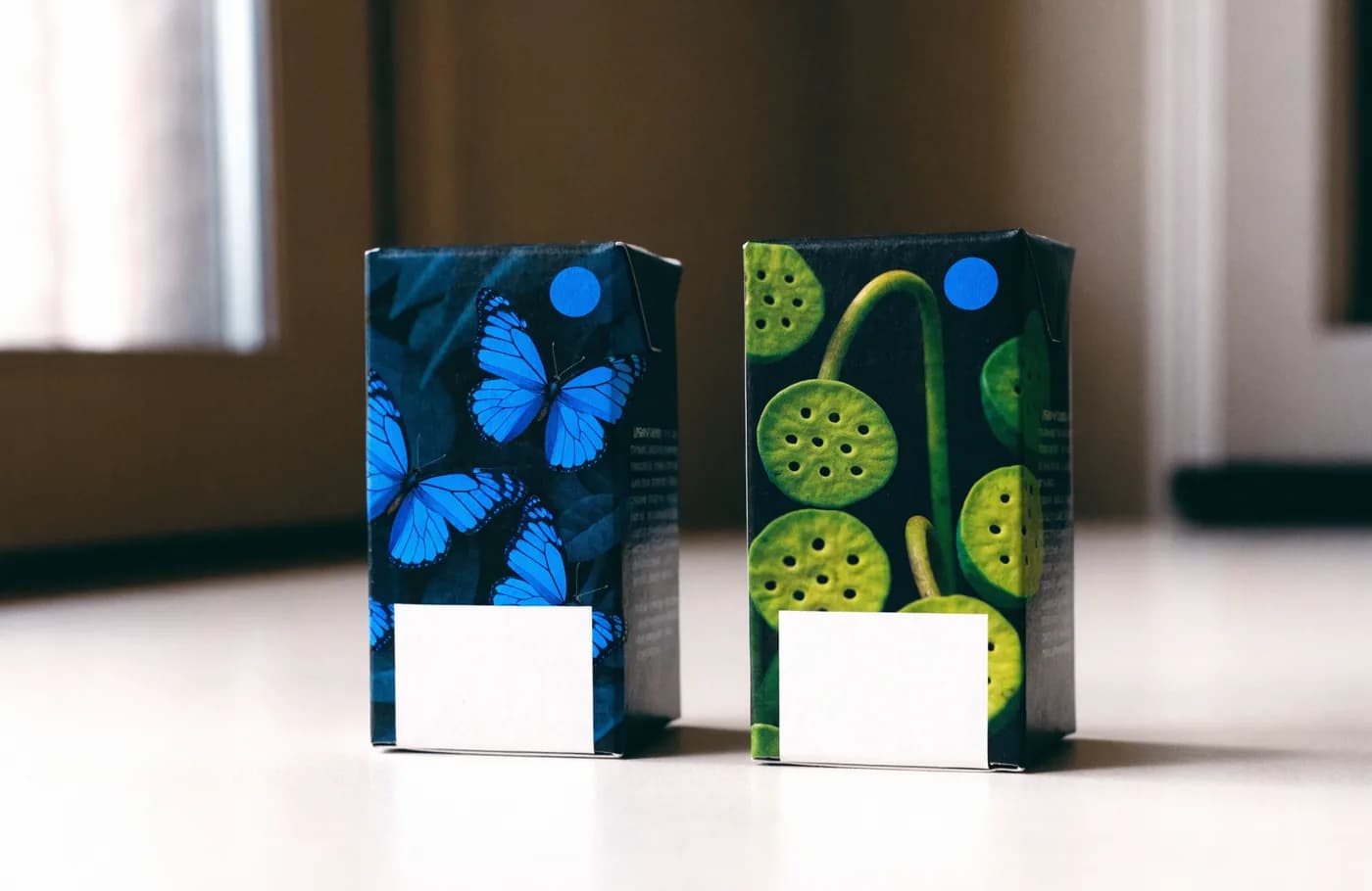 Deux boîtes de cartouches d'encre debout côte à côte, l'une illustrée de papillons bleus, l'autre de motifs végétaux verts